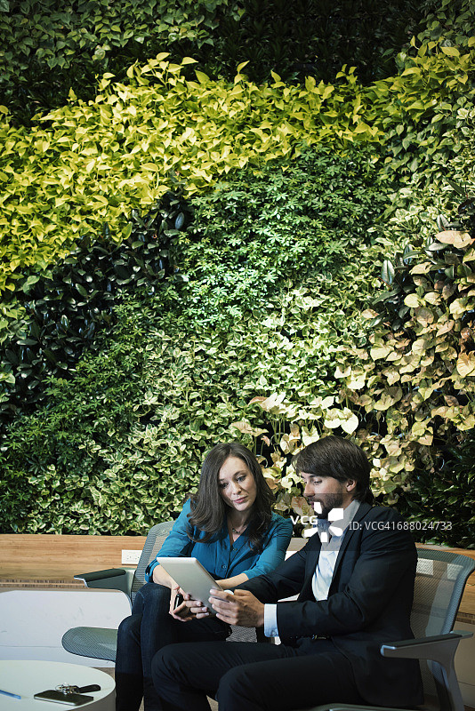 商人和妇女坐在绿色植物墙前，使用数字平板电脑图片素材