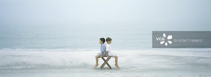 在海滩上，两个男孩背靠背坐在凳子上图片素材