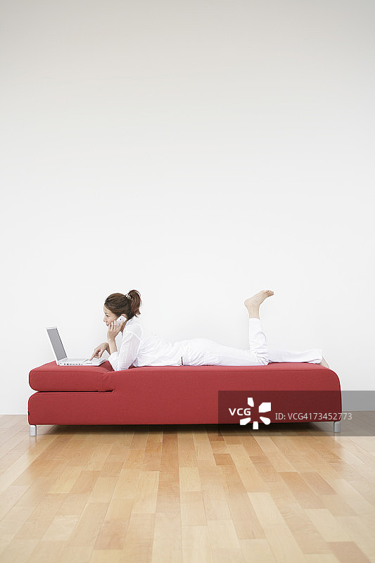 一个年轻女子的侧影躺在沙发上，一边用笔记本电脑一边打手机图片素材