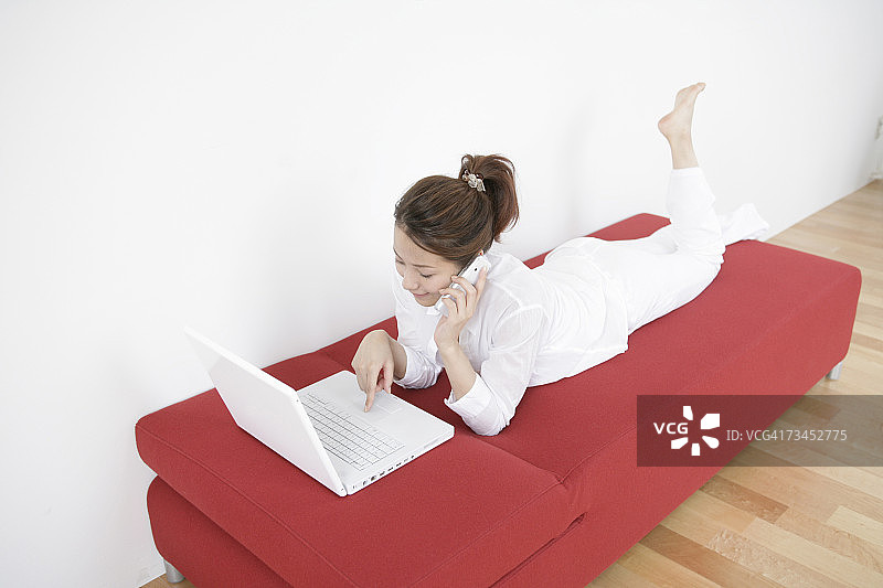 一个年轻女子躺在沙发上使用笔记本电脑，同时在讲手机的高角度视图图片素材
