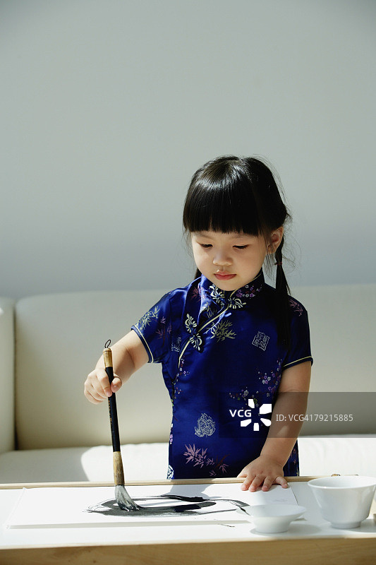 一个穿着蓝色丝绸旗袍的小女孩正在写中国书法图片素材