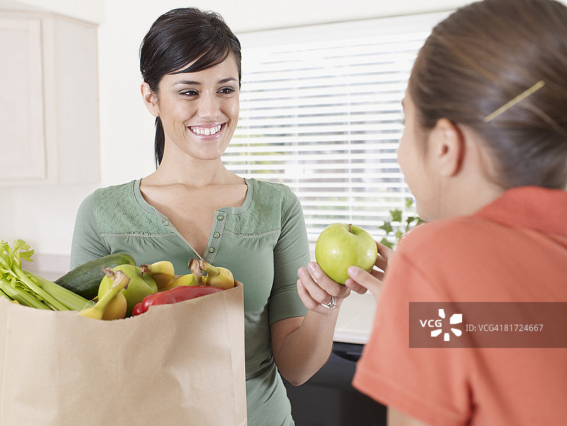 一名妇女微笑着从杂货店的袋子里拿出一个苹果递给厨房里的小女孩图片素材