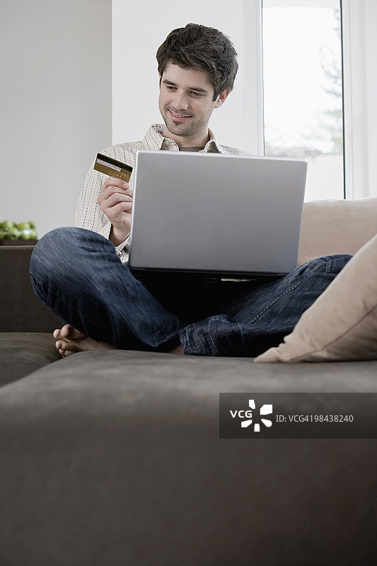 一个年轻人坐在沙发上，用手提电脑拿着信用卡图片素材