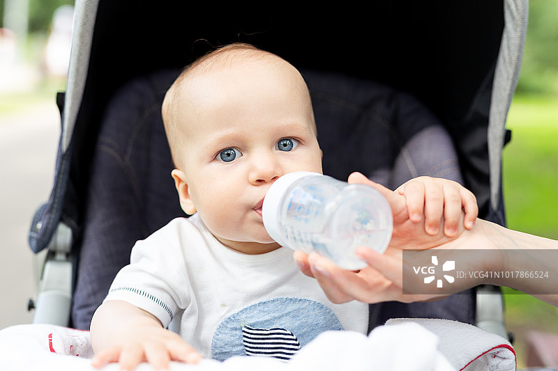 在炎热的夏天，小孩子坐在婴儿车里，喝着瓶子里的水。妈妈给男婴喂奶。在阳光直射下有脱水危险。儿童健康的生活方式图片素材