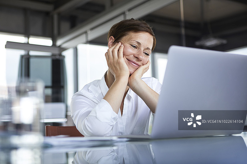 微笑的女商人坐在玻璃桌子在办公室看着笔记本电脑图片素材