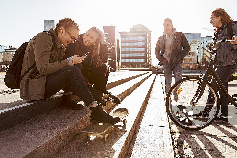 在城市里，当朋友们站在自行车和滑板上时，十几岁的女孩们在台阶上聊天图片素材