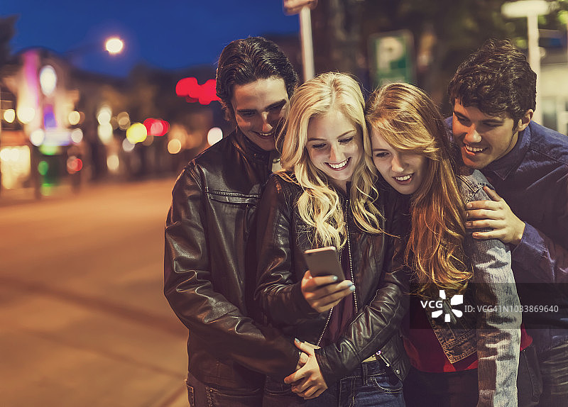 四个朋友挤在人行道上看一部智能手机，屏幕的光芒照亮了他们的脸图片素材