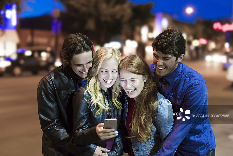 四个朋友聚在人行道上看智能手机，屏幕发出的光芒照亮了他们的脸，他们笑了起来图片素材