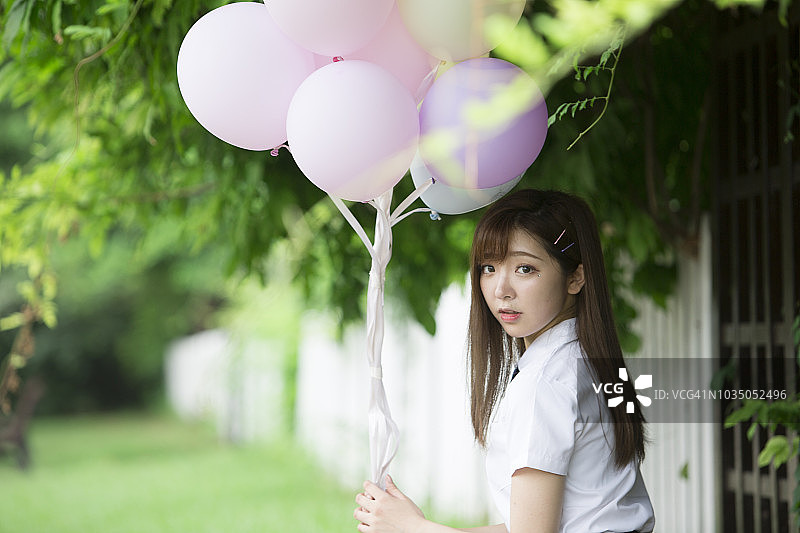 夏天五颜六色的气球。年轻的成年亚洲女孩的肖像抱着一束彩色气球在绿树的背景图片素材