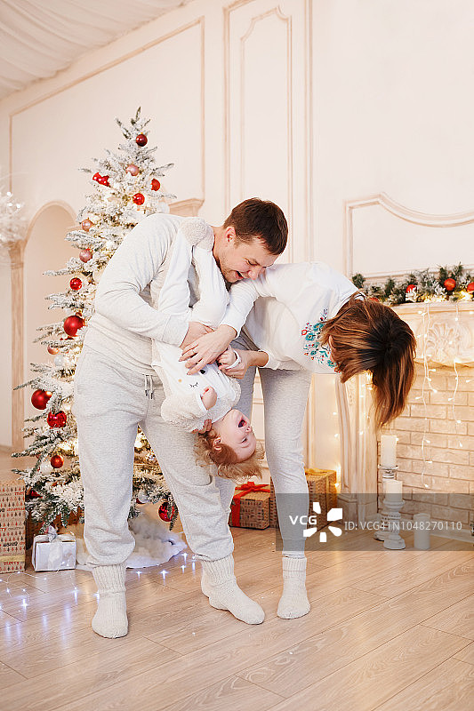 一个快乐的年轻的家庭摇着他们的小女儿微笑着，面对着美丽的室内灯光，有一棵圣诞树和一个白色的壁炉。圣诞节的全家福。宝贝看镜头图片素材