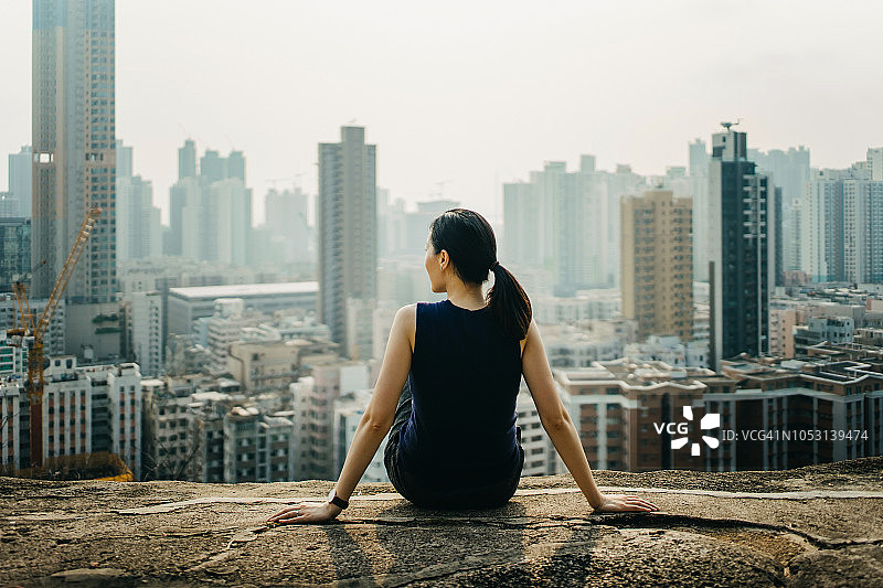 一名年轻女子坐在山顶上，欣赏着壮观的城市天际线的全景图片素材