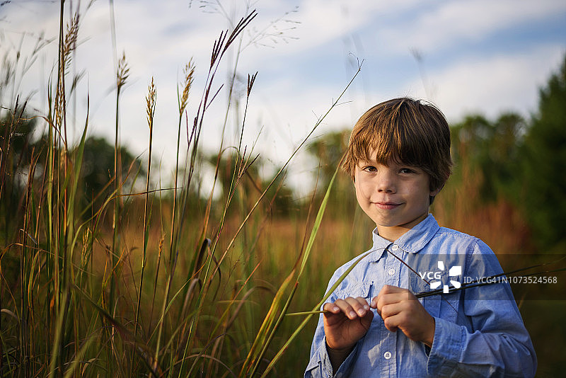 一个微笑的男孩的肖像站在田野在日落采摘长草，美国图片素材