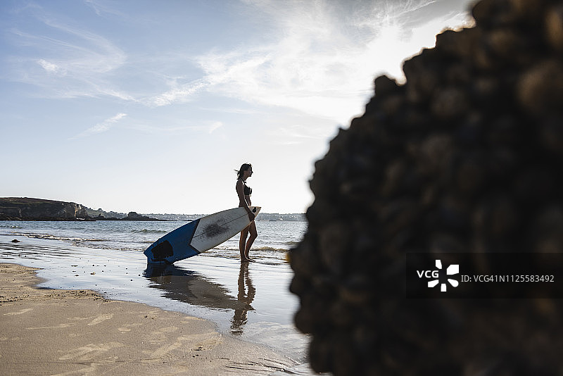 法国，布列塔尼，一个年轻女子带着冲浪板站在海边图片素材