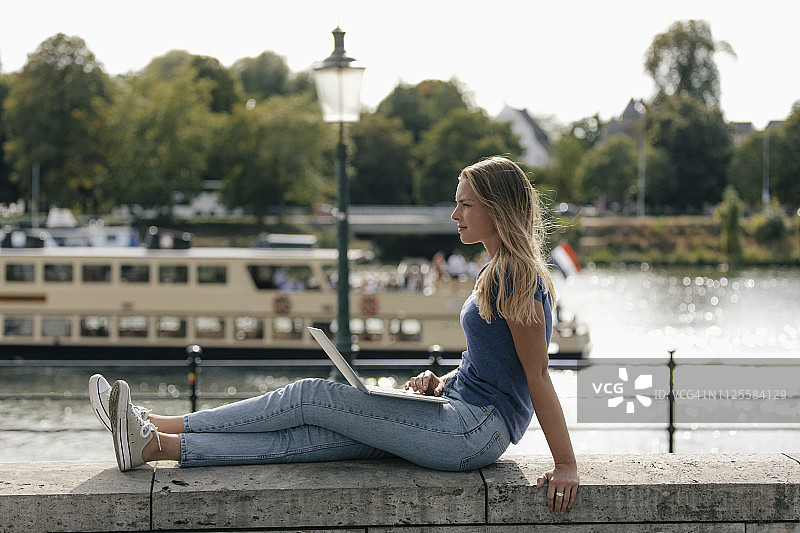 荷兰，马斯特里赫特，一名年轻女子坐在河边的一面墙上，手提电脑图片素材