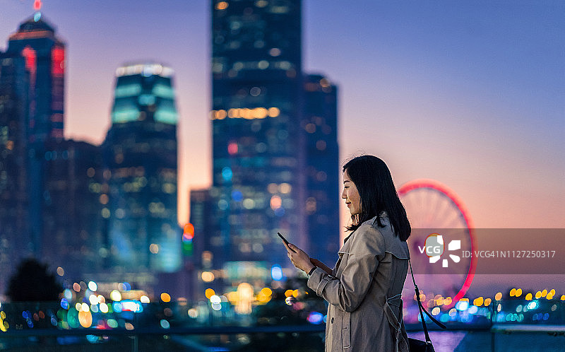 自信的年轻女商人用智能手机对抗香港金融区的现代摩天大楼景观图片素材