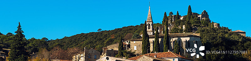 山坡建筑屋顶的教堂在蓝天之上。La Roque-sur-Ceze公社，位于法国南部加尔省图片素材