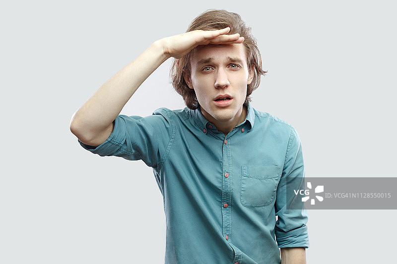 肖像细心英俊的长头发的金发青年男子在蓝色休闲衬衫站在前额，手在看相机。图片素材