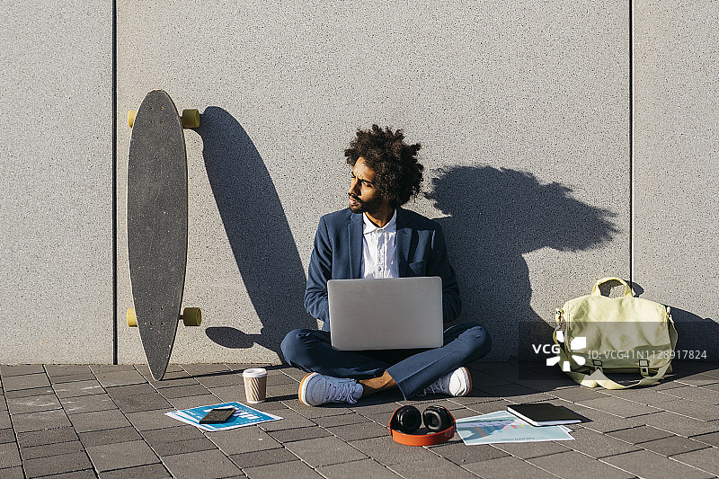 年轻的商人坐在户外的墙上用笔记本电脑工作图片素材