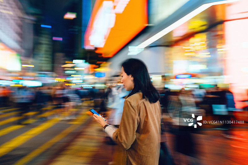 一名忙碌的女性在香港市中心的街道上使用智能手机的模糊动作，背景是人群和明亮的城市场景图片素材