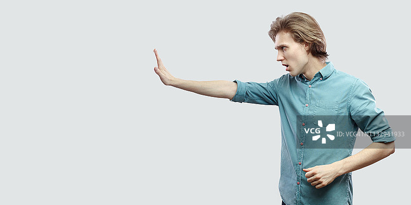 侧视图肖像严肃英俊的长头发金发青年男子在蓝色休闲衬衫站与停止手势手势。图片素材