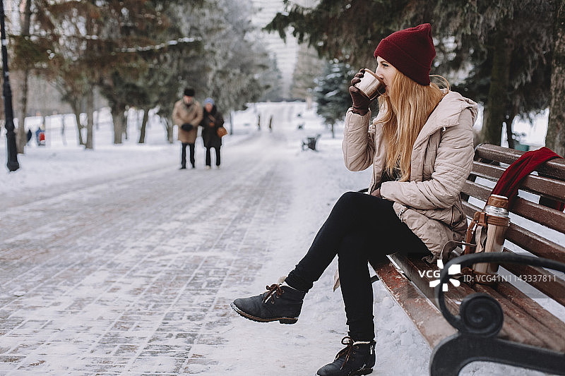 一个女人在冬天坐在公园靠树的长椅上喝咖啡图片素材