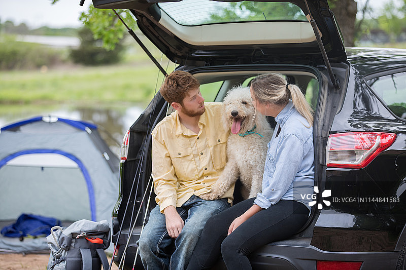 快乐的年轻夫妇坐在汽车与狗露营旅行图片素材