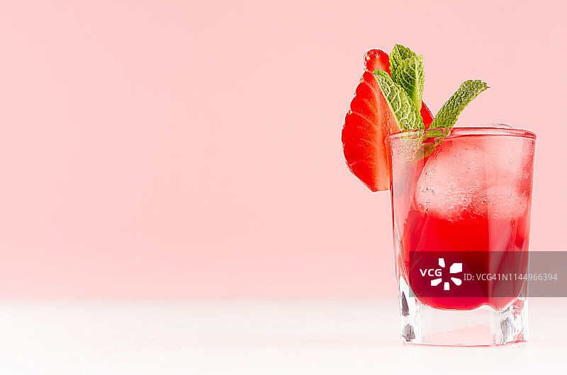 明亮的红色酒精饮料在小酒杯，草莓片，冰块，绿色薄荷在时尚的现代酒吧室内柔和的粉红色。图片素材