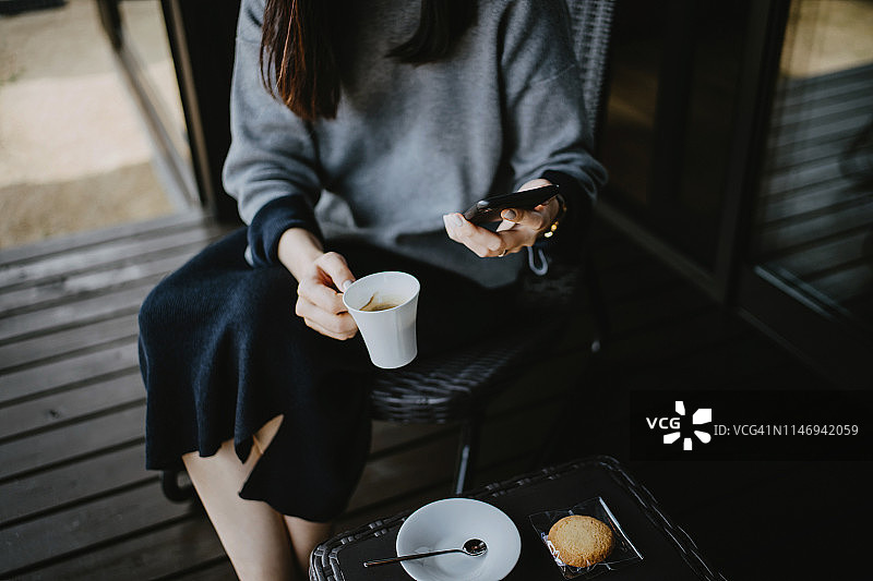近距离的年轻亚洲女子使用智能手机，而放松与咖啡和饼干在阳台图片素材