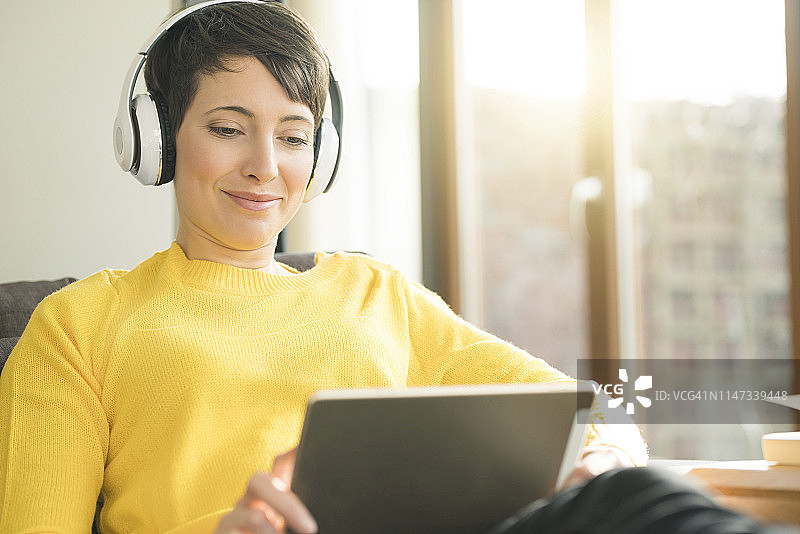 一幅快乐女人戴着耳机坐在家里的沙发上看数字平板电脑的肖像图片素材