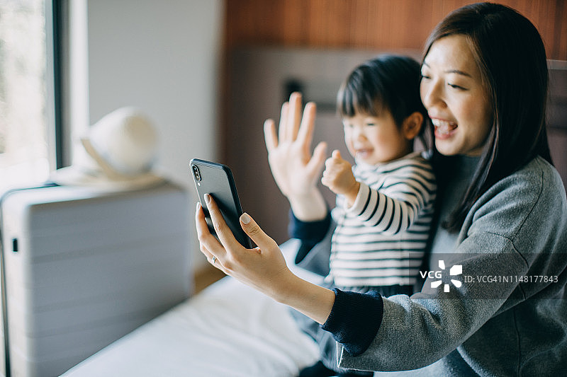 年轻的亚洲母亲和可爱的小女儿有智能手机视频电话与家人在酒店房间度假和愉快的微笑图片素材