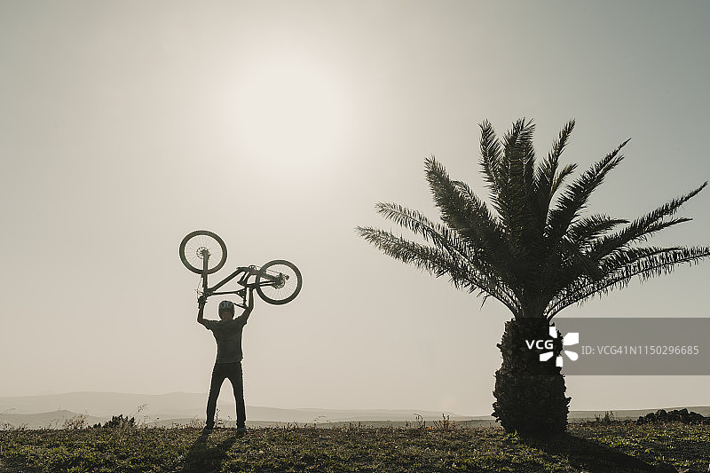 西班牙，兰萨罗特，登山运动员在旅行中举起他的自行车旁边的棕榈树图片素材