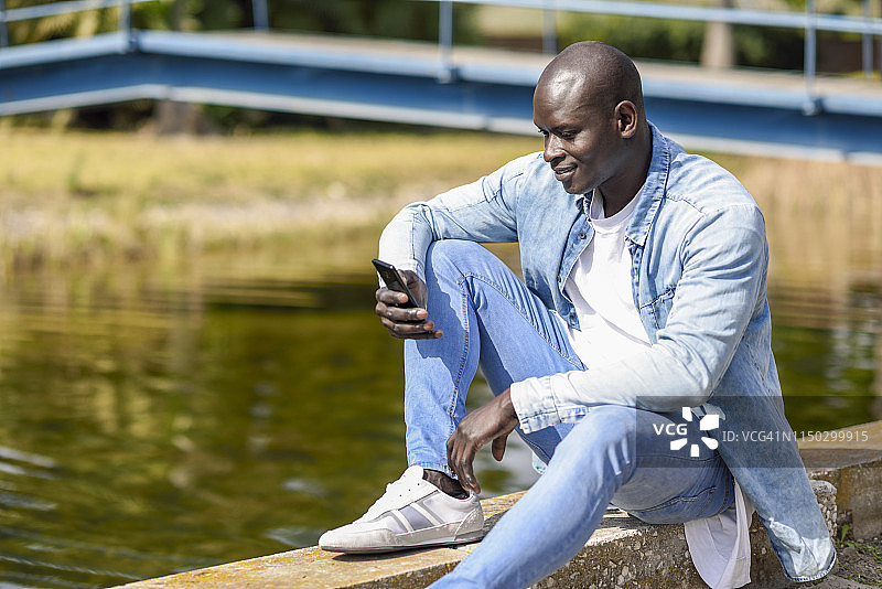 穿着休闲牛仔服装的男子坐在河边玩智能手机图片素材