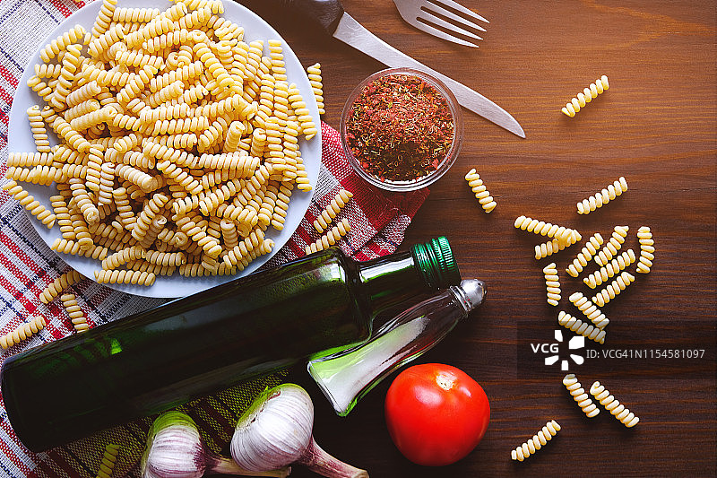 在一张深色的木桌上放着意大利面、橄榄油、香料、西红柿、盐、大蒜和刀叉。顶部视图与复制空间。意大利菜的概念。烹饪素食或纯素食物。图片素材