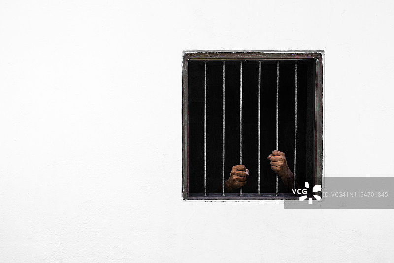 囚犯的双手放在生锈的铁条窗户和牢房白色的水泥墙上图片素材