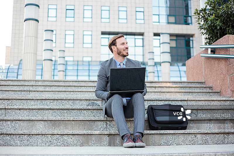 年轻英俊的商人在办公大楼前用笔记本电脑工作。低角度的观点图片素材
