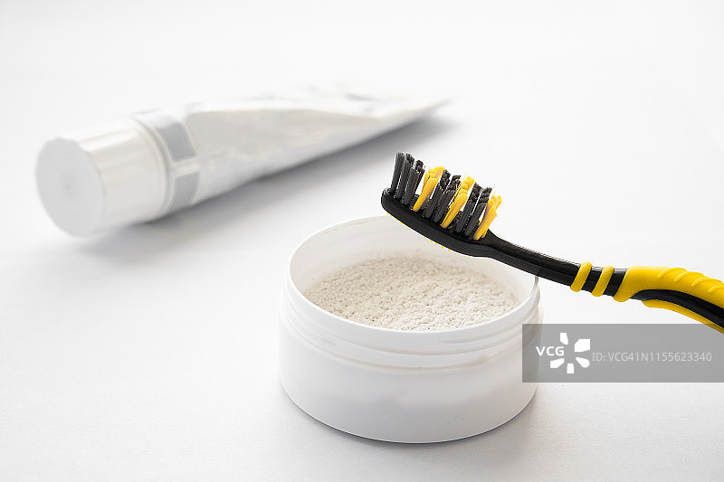 黄黑相间的牙刷放在一罐牙粉上，背景是白色背景上的一管牙膏。口腔护理和牙齿美白的理念。图片素材