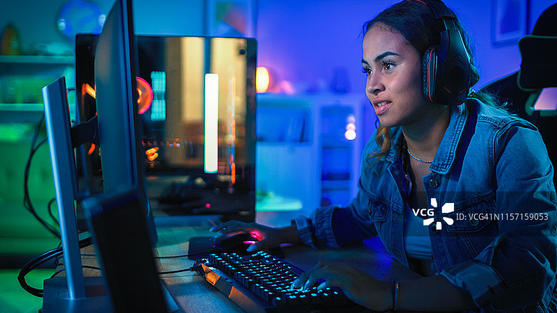 戴着耳机的漂亮和兴奋的黑人博主女孩正在她的电脑上玩第一人称射击在线视频游戏。房间和电脑都有彩色霓虹灯。温馨的家庭之夜。图片素材