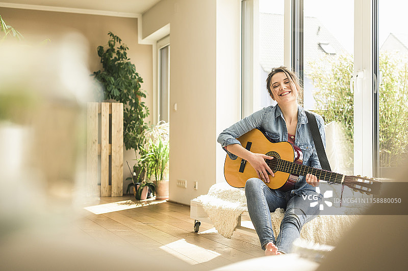 快乐的女人坐在家里的窗边弹吉他图片素材