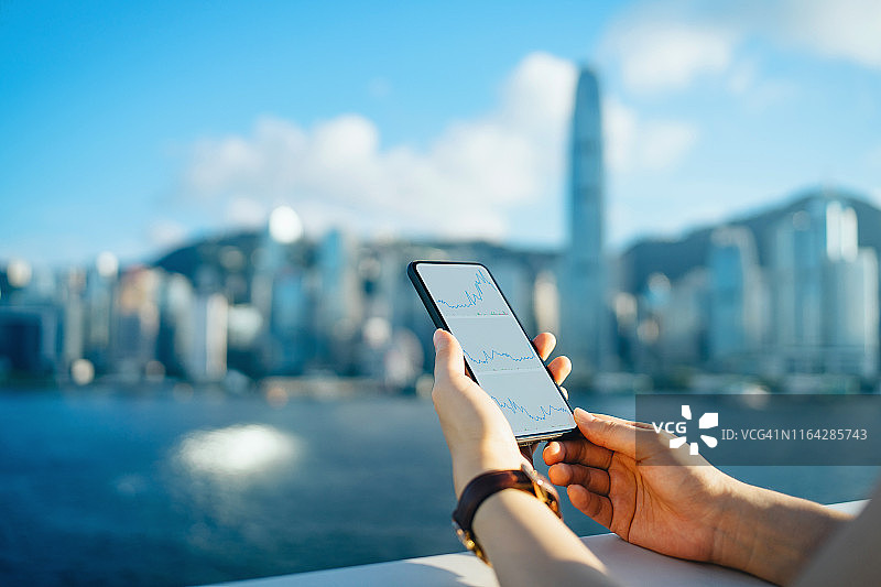 在一个阳光明媚的日子里，一名女子在维多利亚港海滨漫步，在香港标志性城市天际线的映衬下，用智能手机阅读金融交易数据图片素材