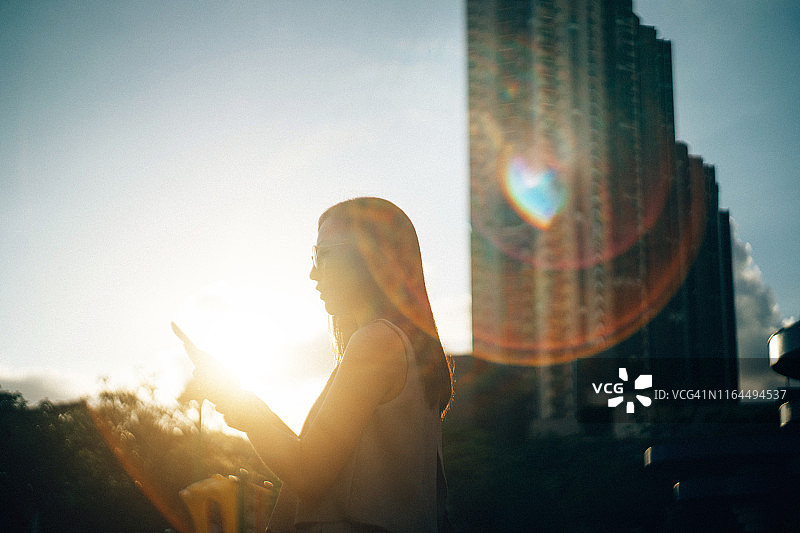年轻女子在美丽的夕阳下使用智能手机，镜头光晕图片素材