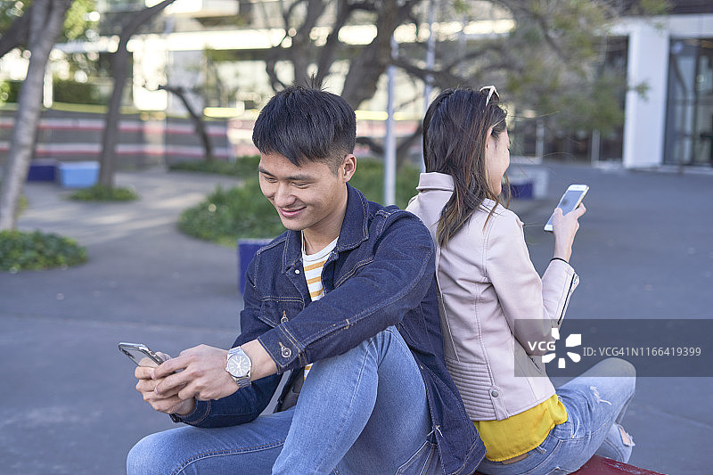 西班牙巴塞罗那，一对轻松的情侣背靠背坐着玩手机图片素材
