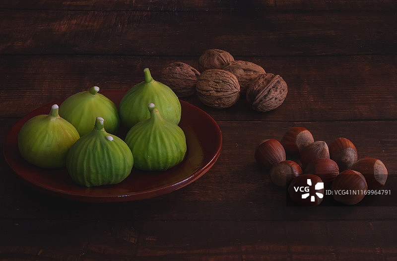 秋天的水果:一组新鲜采摘的绿色无花果放在粘土盘子里，坚果和榛子放在旧的深色木桌上图片素材