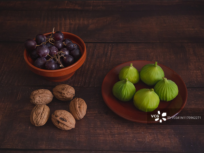 秋天的水果:黑色的葡萄放在粘土碗里，新鲜收获的绿色无花果放在粘土盘子里，坚果放在旧的深色木桌上图片素材