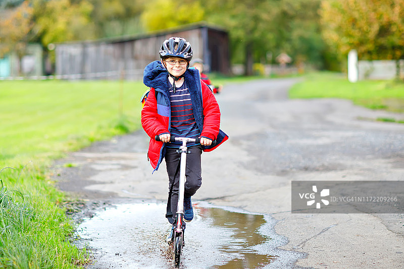 可爱的小男孩骑着滑板车在往返学校的路上。7岁的小男生开车穿过雨坑。有趣的快乐的孩子在五颜六色的时装和头盔。图片素材