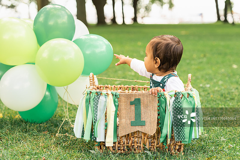 可爱的婴儿坐在一个编织的篮子里，气球和绿色缎带横幅上写着“一个”。一个奇思妙想的热气球即将在美丽的森林空地上起飞。图片素材