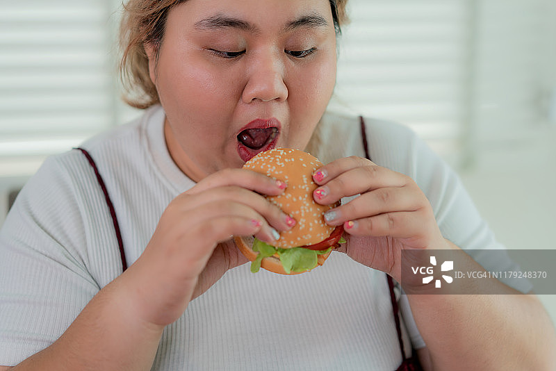 亚洲超重妇女咬快餐汉堡特写，不健康的营养和暴吃。她吃垃圾食品看起来很开心。饮食不健康的生活方式。图片素材