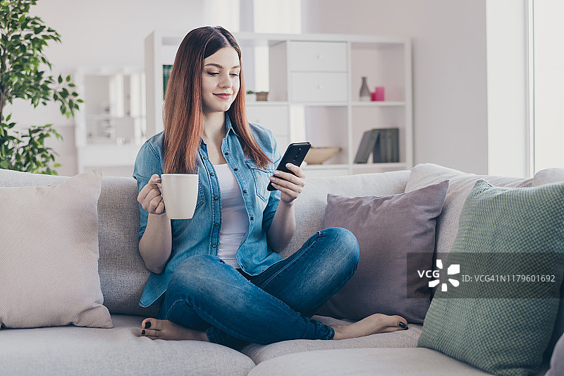 照片惊人的女士电话和热饮料在手写社交网络博客坐在舒适的沙发穿着牛仔裤衣服公寓室内的新帖子图片素材