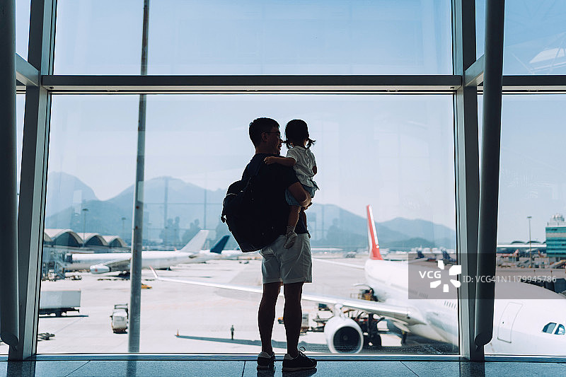 在机场等待起飞的时候，快乐的年轻亚洲父亲抱着可爱的小女儿，透过窗户看着飞机的剪影图片素材