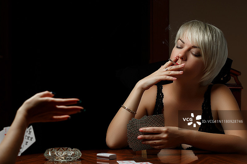 一个年轻的白人金发女郎在黑色内衣坐在桌子和打牌与另一个女人脱衣和吸烟。图片素材