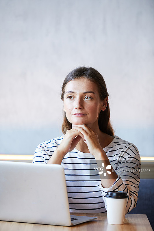 严肃沉思的女性网络营销者，穿着休闲服装坐在咖啡馆的桌子上，倚着头思考网页设计图片素材
